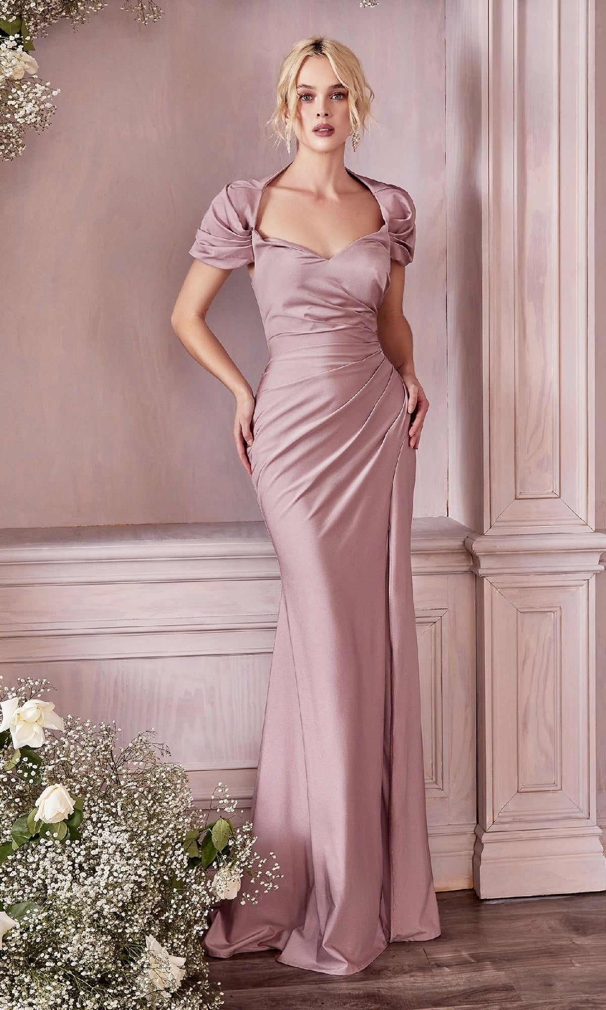 A woman stands indoors surrounded by light wood paneling and floral arrangements, wearing the CLKV1061 Cap-Sleeve Long Satin Prom Dress—a floor-length, fitted mauve gown featuring cap sleeves and a sweetheart neckline.